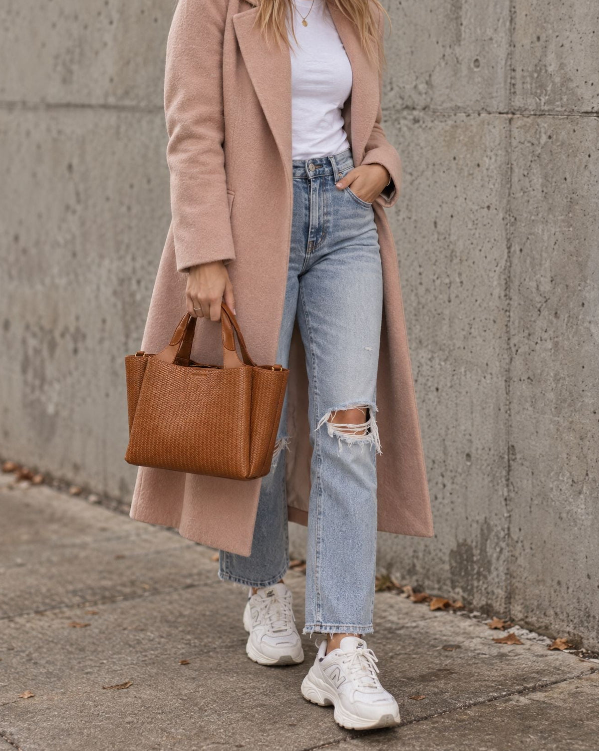 A woman stands against a concrete wall wearing a long blush pink coat over a white top and light blue ripped jeans, carrying a brown leather tote bag.