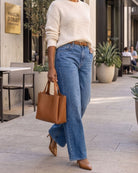 A woman walking outdoors in a modern urban setting, wearing a cream ribbed knit sweater, high-waisted blue wide-leg jeans, a brown belt, tan pointed-toe heels, and carrying a structured tan leather tote bag.